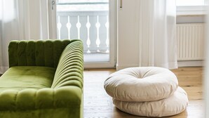 Een kamer met een groene bank, een witte poef en uitzicht door een glazen deur op een balkon.