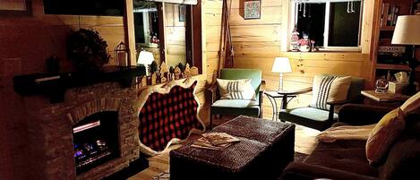A cozy log cabin interior with a fireplace, a green armchair, a small table with glasses, and a window with a view of the outside.