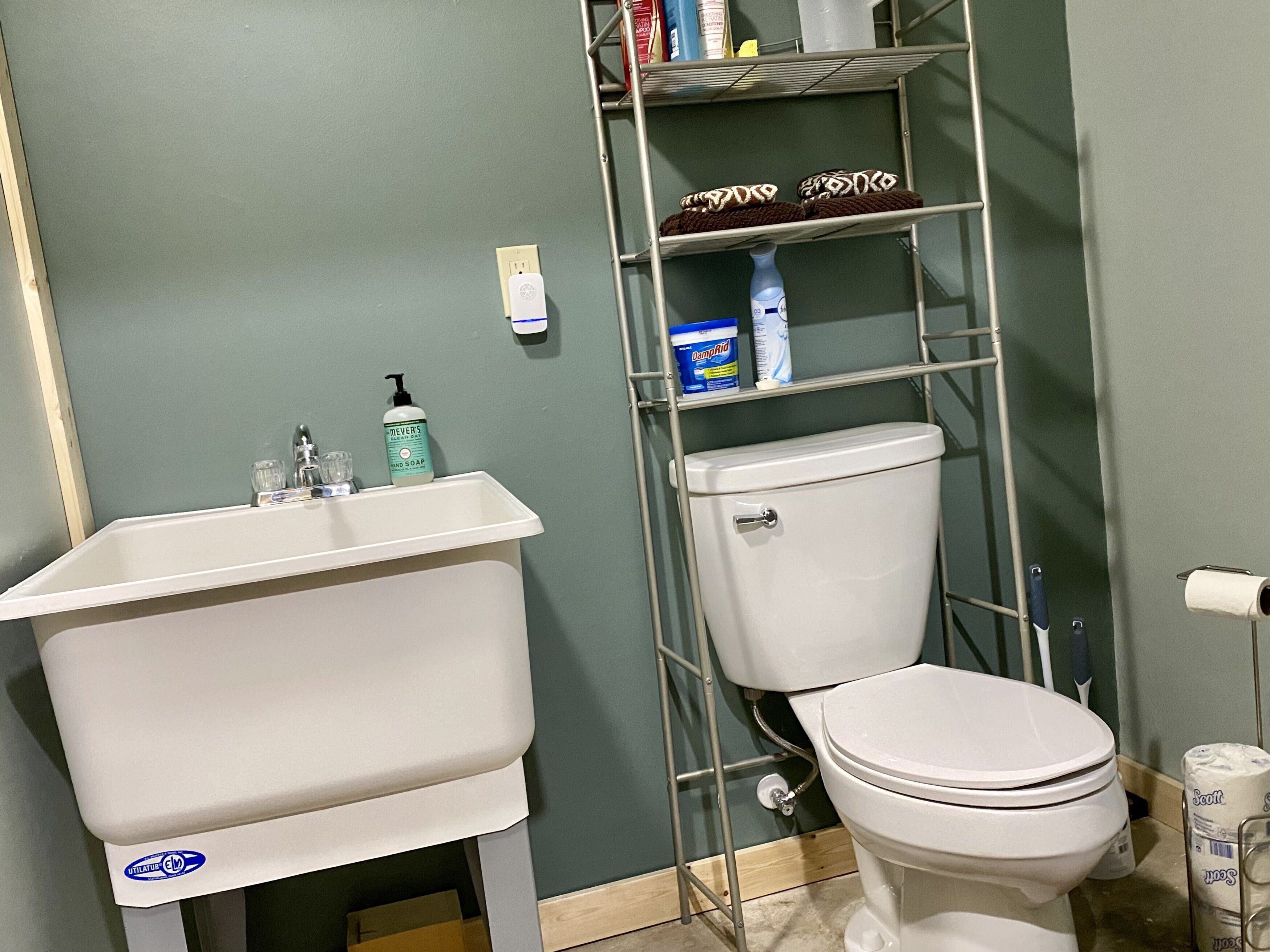 A bathroom with a white sink, toilet, and a metal shelf above the toilet holding cleaning supplies.