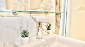 Un baño con pared de mármol, repisa de vidrio con vasos, lavamanos con grifo y planta en maceta.