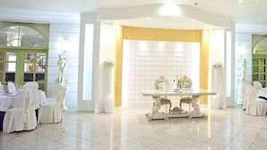Un salón amplio con una mesa central y sillas, rodeado de sillas con fundas blancas y grandes columnas.