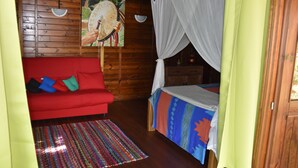 A wooden cabin room with a red sofa, a bed with a canopy, a colorful rug, and a painting on the wall. - CHALET "Danse du Tambour" 2 people - (Pointe-Noire)