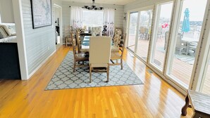 Ein geräumiges Zimmer mit Holzboden, einem Esstisch mit Stühlen, einer Kandelaber und großen Fenstern mit Blick ins Freie.