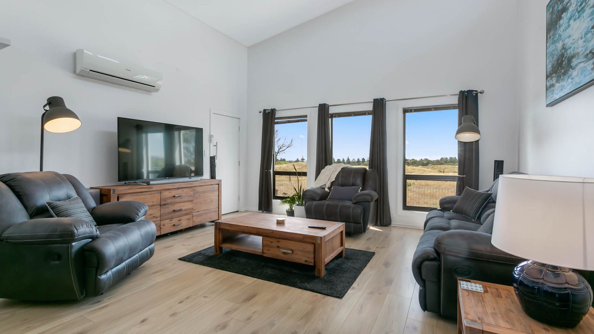 A modern living room with a flat-screen TV, leather sofas, a wooden coffee table, and large windows offering a view of the outdoors.