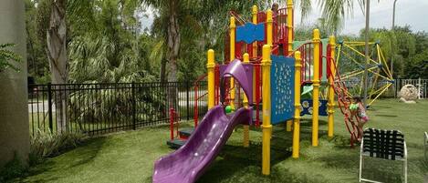 Un parque infantil con un tobogán morado, estructuras de juego amarillas y rojas, y un banco blanco y negro.