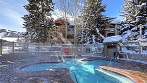 Un espace piscine avec un jacuzzi, entouré d’arbres et de bâtiments couverts de neige.