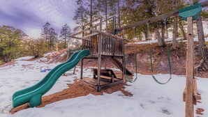 Een houten speelconstructie met een groene glijbaan en schommels, omringd door bomen en sneeuw.