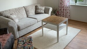Ein Wohnzimmer mit einer Couch, einem Couchtisch, einem gemusterten Hocker, einem Wandbild und einem Fenster mit Vorhängen.