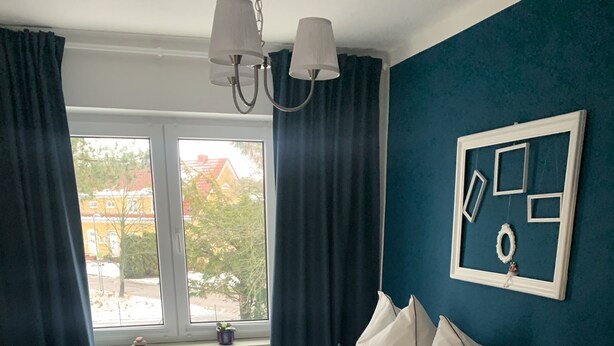 Ein Schlafzimmer mit Bett, Fenster, Vorhängen, einer Wandleuchte und einem gerahmten Bild.