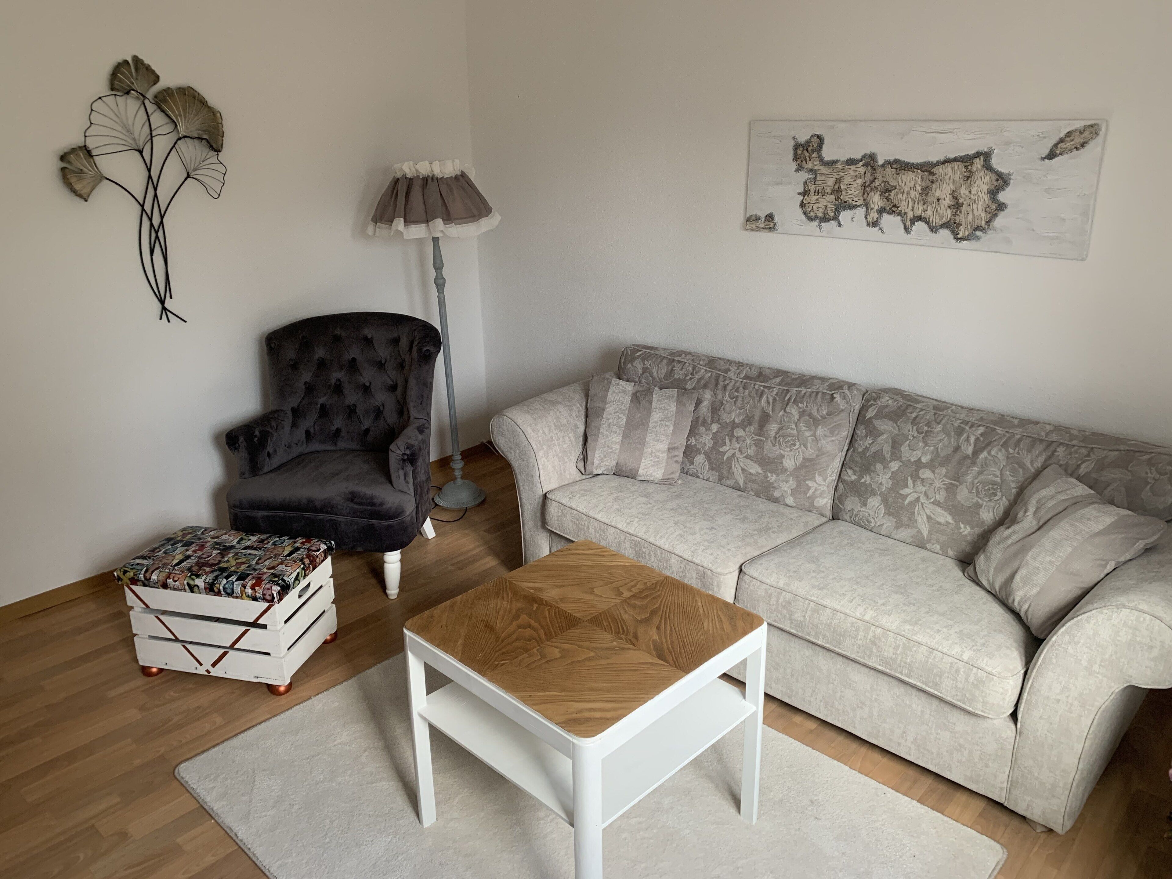 A living room with a dark velvet armchair, a light-colored sofa, a wooden coffee table, a white ottoman, a floor lamp, and wall art.