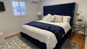 A bedroom with a large bed, a dark blue headboard, a geometric patterned rug, a bedside table with a lamp, and a window with blinds.