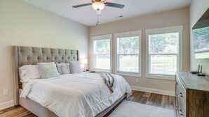 Un dormitorio con cabecera capitonada, cama con ropa de cama blanca, ventilador de techo y tres ventanas con persianas.