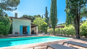 A poolside area with lounge chairs, a table with a bottle and glass, and a view of a building with blue shutters. - La Tropézienne - Welkeys (Gassin)