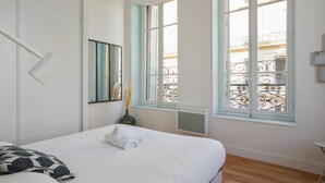 Un dormitorio con cama, una lámpara de noche, un florero con plumas de pampa y una ventana que ofrece vistas a un balcón.