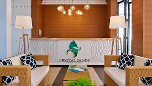 Een receptie met een houten receptiebalie, twee rieten banken met gestreepte kussens, en een logo voor 'Crystal Sands Simply Exotic'.