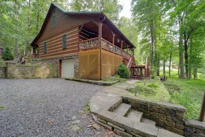 Eine rustikale Holzhütte mit Veranda, Steinstufen und einem Schotterweg, umgeben von Bäumen.