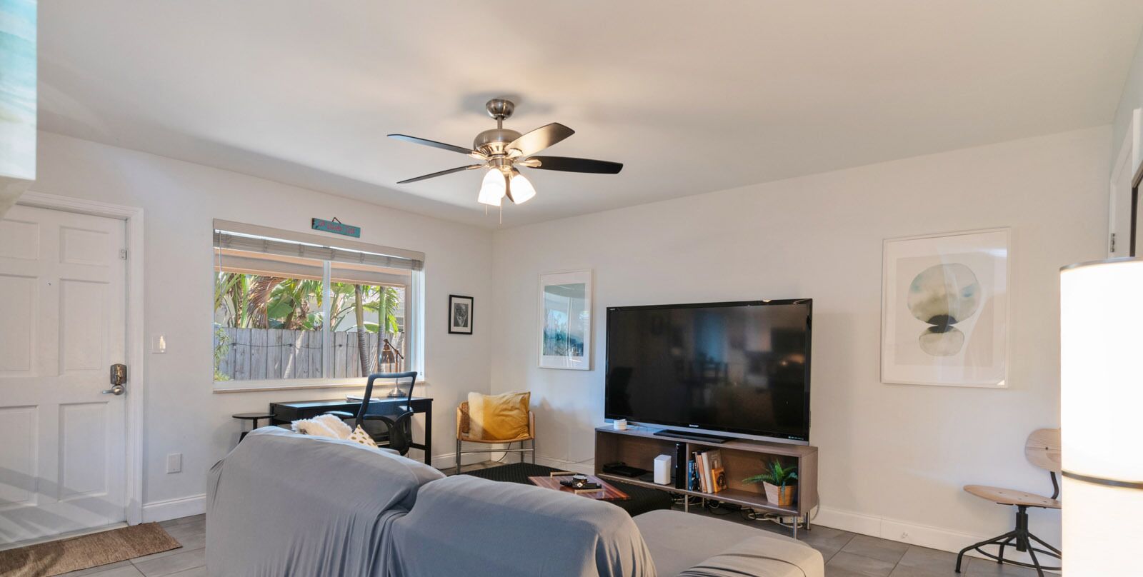 A living room with a ceiling fan, a television, a sofa, a chair, and a window with a view of greenery.