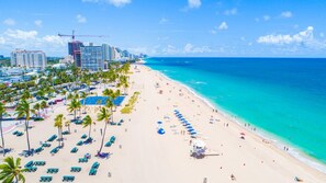 Una playa con filas de palmeras y sillas de playa, un océano azul claro y edificios al fondo.