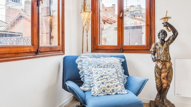 A room with a blue chair, a statue, and a view of a cityscape through large windows. - GuestHost - Bright apartment, of about 80 sqm with period furniture, which can accommodate up to 4 people, located on the third floor (NO lift) of a typical Venetian building. The building boasts a very central position: it is a two-minute walk from Piaz (Venezia)