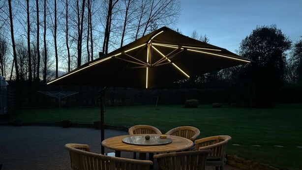 Et udendørs siddeområde med et rundt bord og stole, en stor parasol og en græsplæne i baggrunden.