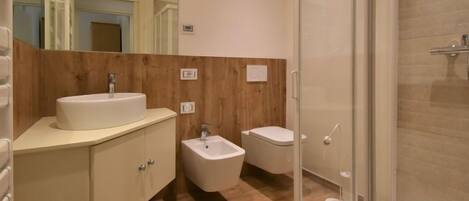 A modern bathroom with a wooden vanity, a small sink, a bidet, and a glass-enclosed shower.