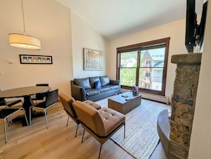Uma sala de estar moderna com sofá, mesa de centro e cadeiras, uma mesa de jantar com cadeiras, lareira e uma grande janela com vista.