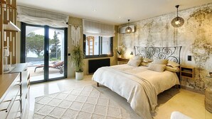 A bedroom with a large bed, a view of the outdoors, a potted plant, and a wooden desk.