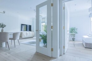 A modern living room with a large white door, a flat-screen TV, a dining area with a white table and chairs, and a small plant on a side table.