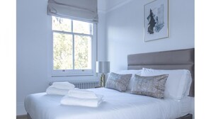 A neatly made bed with white linens and towels, a grey headboard, and a window with a view of trees. - Royal Kensington - Premium 3 bed (London)