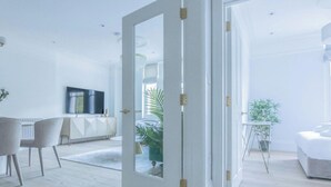 A modern, well-lit room with a large mirror, a dining area with chairs, a sofa, and a small table with a plant. - Royal Kensington - Premium 3 bed (London)