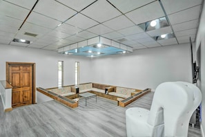 A room with a sectional sofa, a white chair, a wooden door, and a ceiling with recessed lighting.