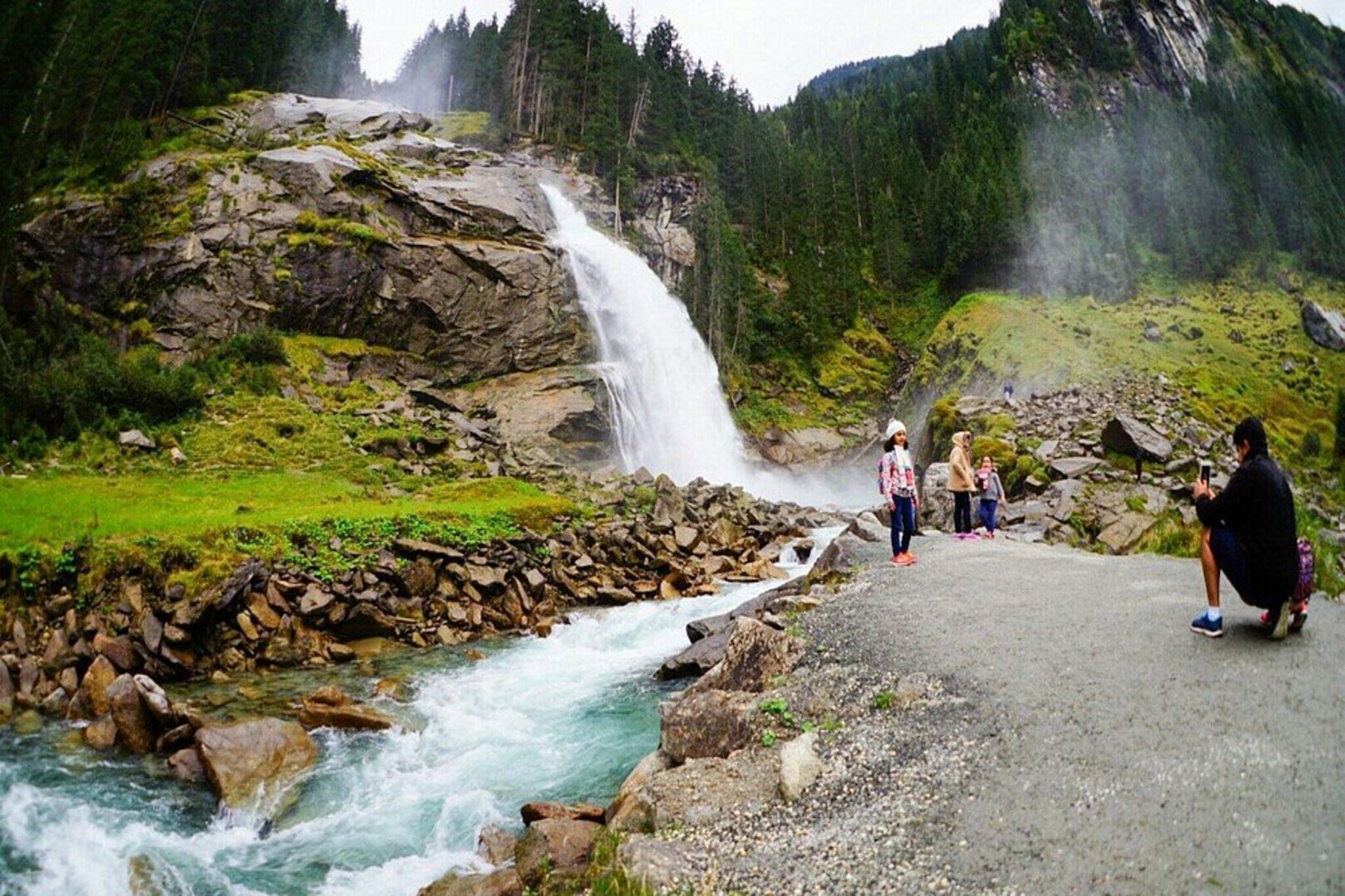 Une cascade d’eau qui dévale des rochers pour se jeter dans une rivière, le tout entouré de verdure et observé par quelques personnes.