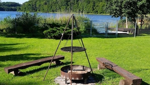 Ein rustikales Grillarrangement im Freien mit Feuerstelle, Picknicktisch und Holzscheiten, umgeben von einer Rasenfläche und Blick auf einen See.