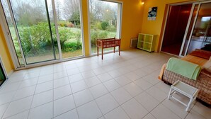 A bright room with large sliding glass doors leading to a garden, a small wooden chair, a wicker sofa, and a green shelf.
