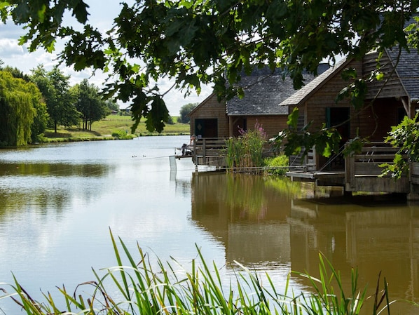 A rustic wooden cabin with a dock extending into a calm lake, surrounded by lush greenery and trees.
