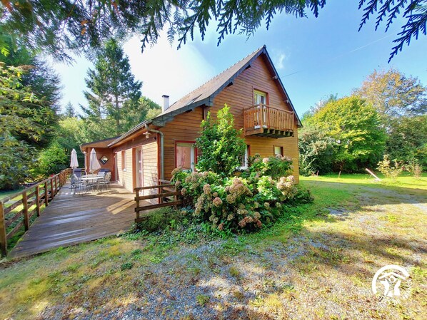 A wooden cabin with a deck, surrounded by greenery and trees.