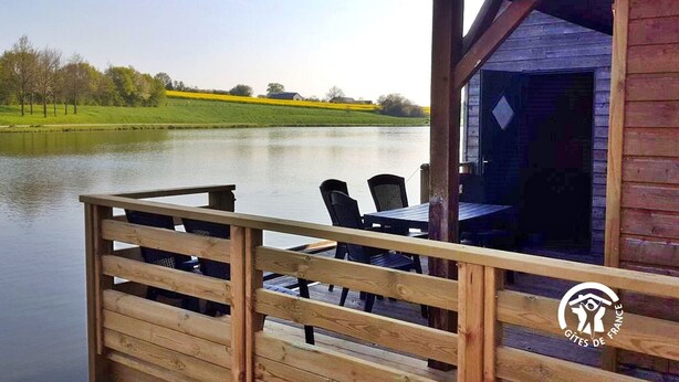 A wooden cabin with a deck overlooking a calm lake, surrounded by greenery.