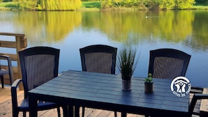 Une terrasse en bois avec une table et des chaises, donnant sur un lac paisible, le tout entouré d’une végétation luxuriante.