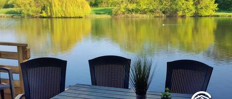 Une terrasse en bois avec une table et des chaises, donnant sur un lac paisible, le tout entouré d’une végétation luxuriante.