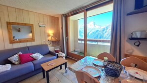 Ein gemütliches Wohnzimmer mit einer Couch, einem kleinen Tisch, einem Esstisch für vier Personen und einem großen Fenster mit Blick auf die Berge.
