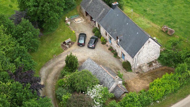 Une vue aérienne d’un bâtiment en pierre, doté d’un toit en pente, entouré d’une végétation luxuriante et bordé d’une allée gravillonnée.