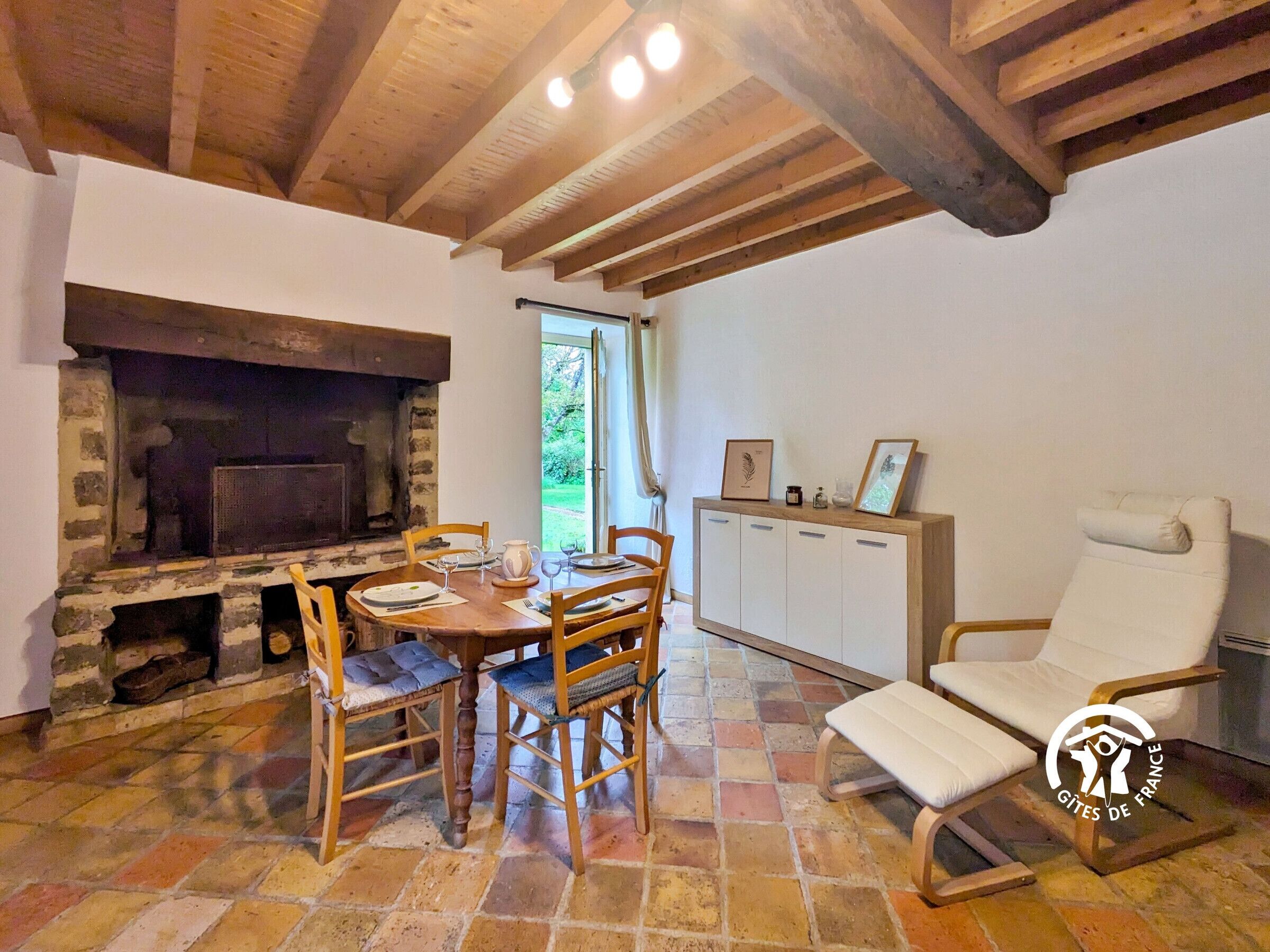 Un intérieur rustique avec une cheminée en pierre, une table à manger en bois et un fauteuil blanc.