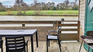 Una terraza de madera con una mesa y sillas, con vistas a un río y campos verdes.