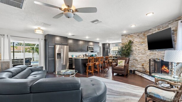 A modern living room with a leather sectional sofa, a dining area with wooden chairs, a kitchen with dark cabinets, and a fireplace. - Quiet, Centrally Located Home w/ Heated Pool, Gas Grill & Private Backyard - Poolside Paradise (Pinellas Park)