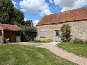 A rustic stone building with a tiled roof, a gravel path leading to a wooden door, and a small seating area with a bench.