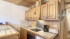 A compact kitchen with wooden cabinets, a stainless steel refrigerator, a microwave, a coffee maker, and a clock on the wall.