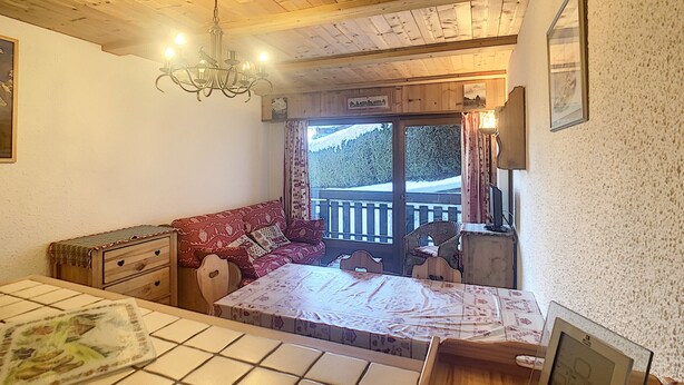 A cozy room with a wooden ceiling, a red sofa, a wooden chest of drawers, a tiled table, and a view of a snowy landscape through a window.
