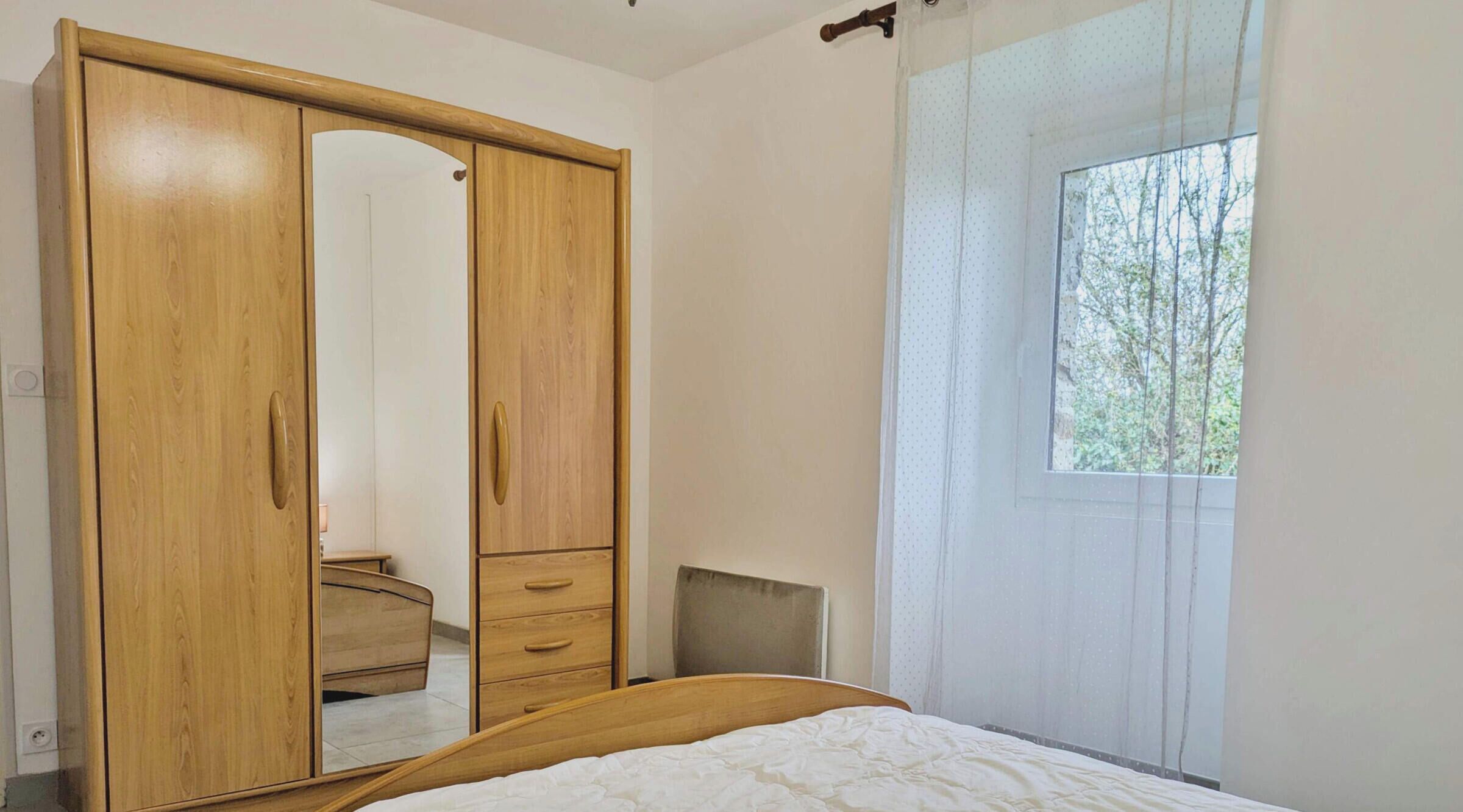 A bedroom with a wooden wardrobe, a bed, a chair, and a window with a view of trees.