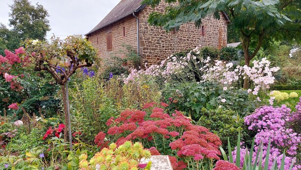 Een stenen gebouw met rieten dak, omgeven door een tuin met verschillende bloeiende planten en struiken.