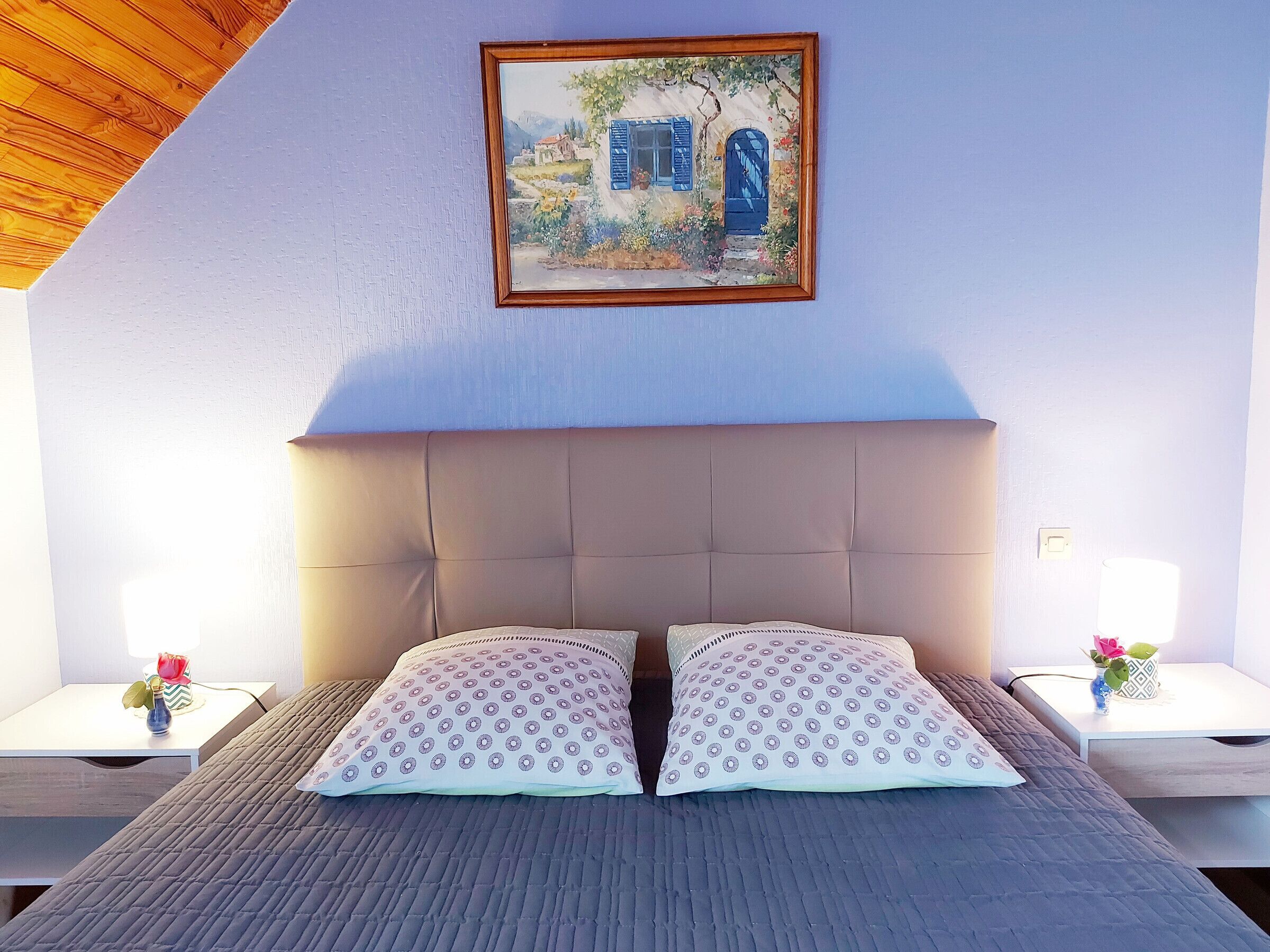 A bedroom with a large bed, two nightstands, a painting on the wall, and a wooden sloped ceiling.